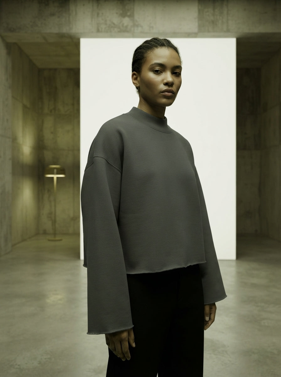 A model wearing a charcoal gray boxy mock neck sweatshirt with raw edges in a minimalist studio setting.
